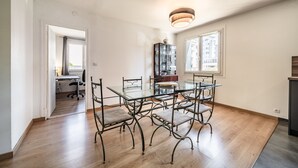 Een eetruimte met een glazen tafel en metalen stoelen, een houten vloer, een keukenhoek en een uitzicht door een raam.