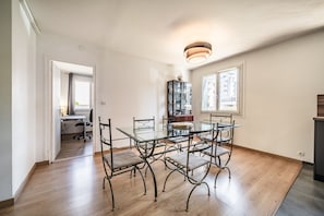 Comedor con mesa de vidrio, sillas de metal, piso de madera, zona de cocina y vistas por una ventana.