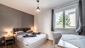 Een slaapkamer met een bed, nachtkastje, lamp, stoel en een raam met gordijnen.