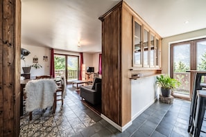 A spacious room with a large wooden cabinet, a dining area with a patterned rug, and a view of greenery outside.