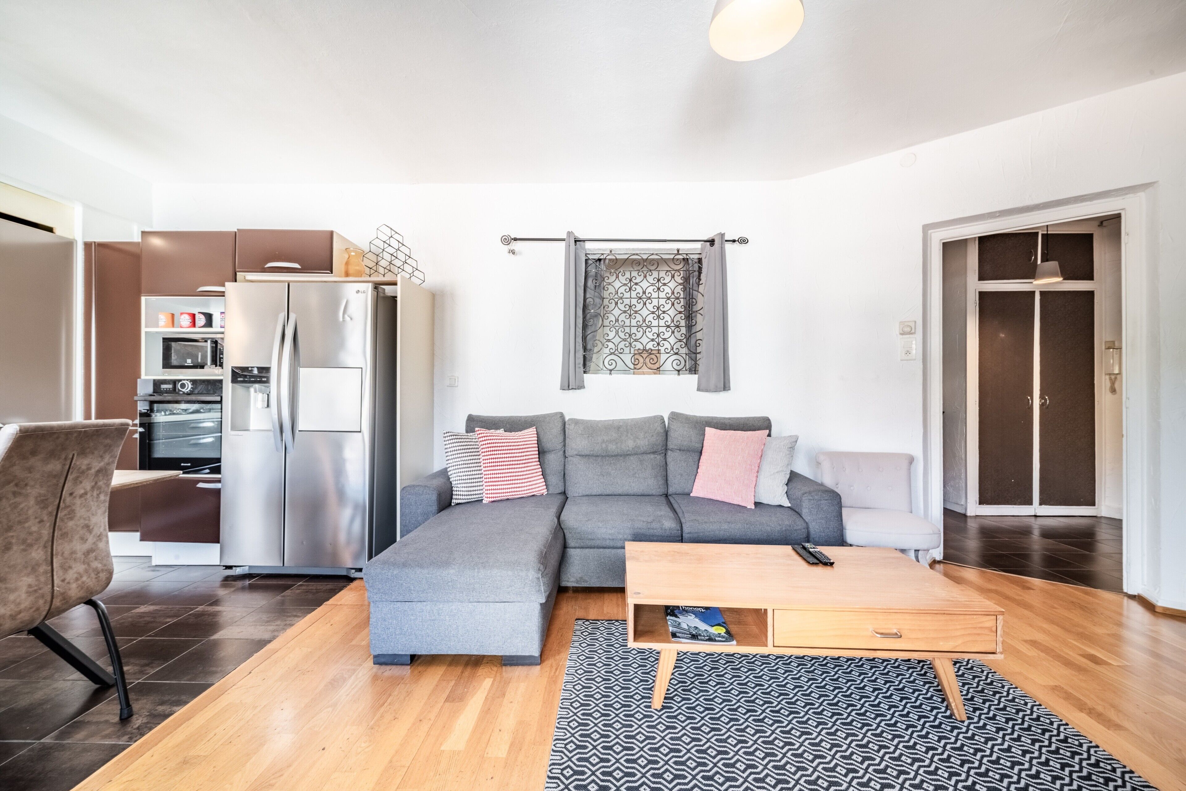 Una sala moderna con un sofá seccional gris, una mesa de centro de madera, una alfombra estampada y una zona de cocina con refrigerador y microondas.