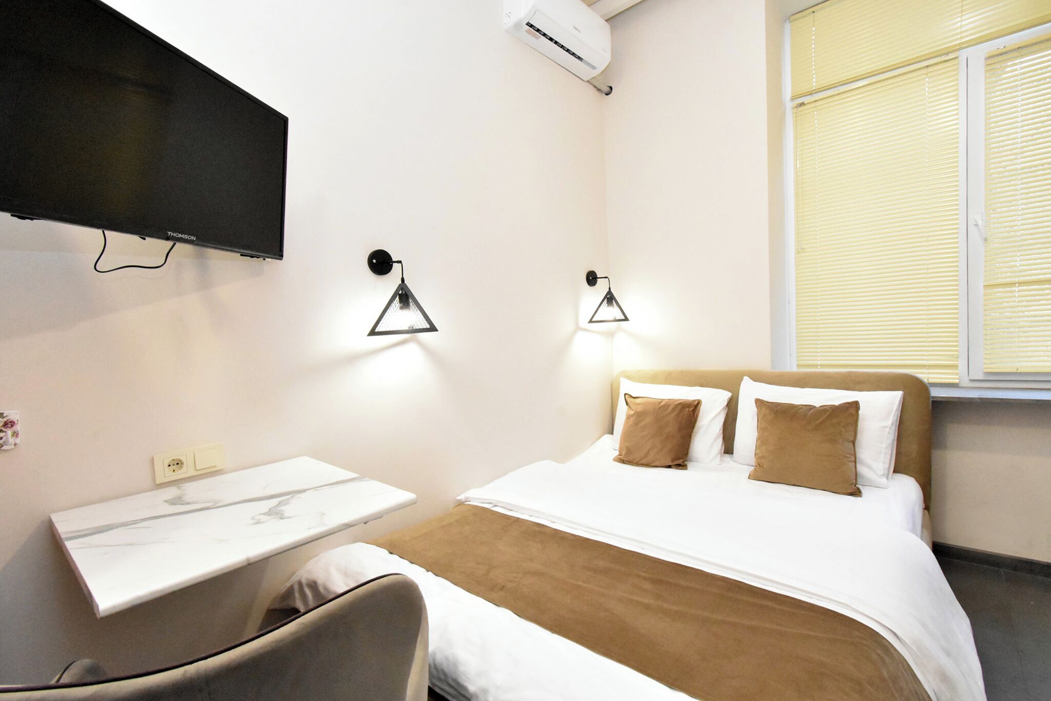 A hotel room with a bed, a desk, a chair, a flat-screen TV, and two wall-mounted lamps.