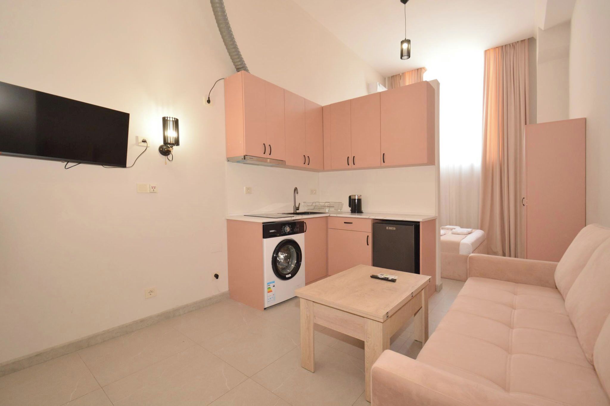 Een compacte woonruimte met een kitchenette, een wasmachine, een bank en een aan de muur bevestigde televisie.