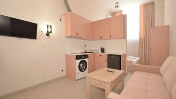 Een compacte woonruimte met een kitchenette, een wasmachine, een bank en een aan de muur bevestigde televisie.