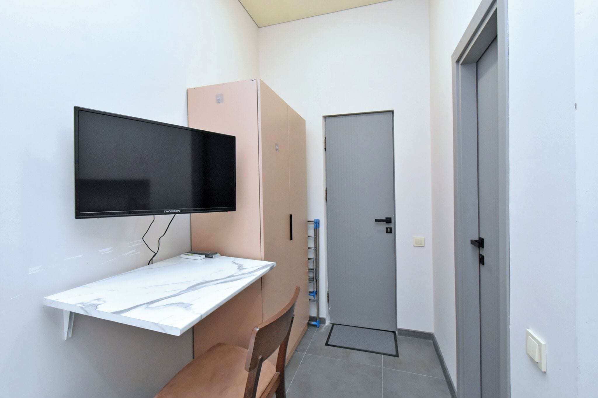 A small room with a flat-screen TV mounted on the wall, a white marble desk, a brown chair, a closed door, and a water dispenser.
