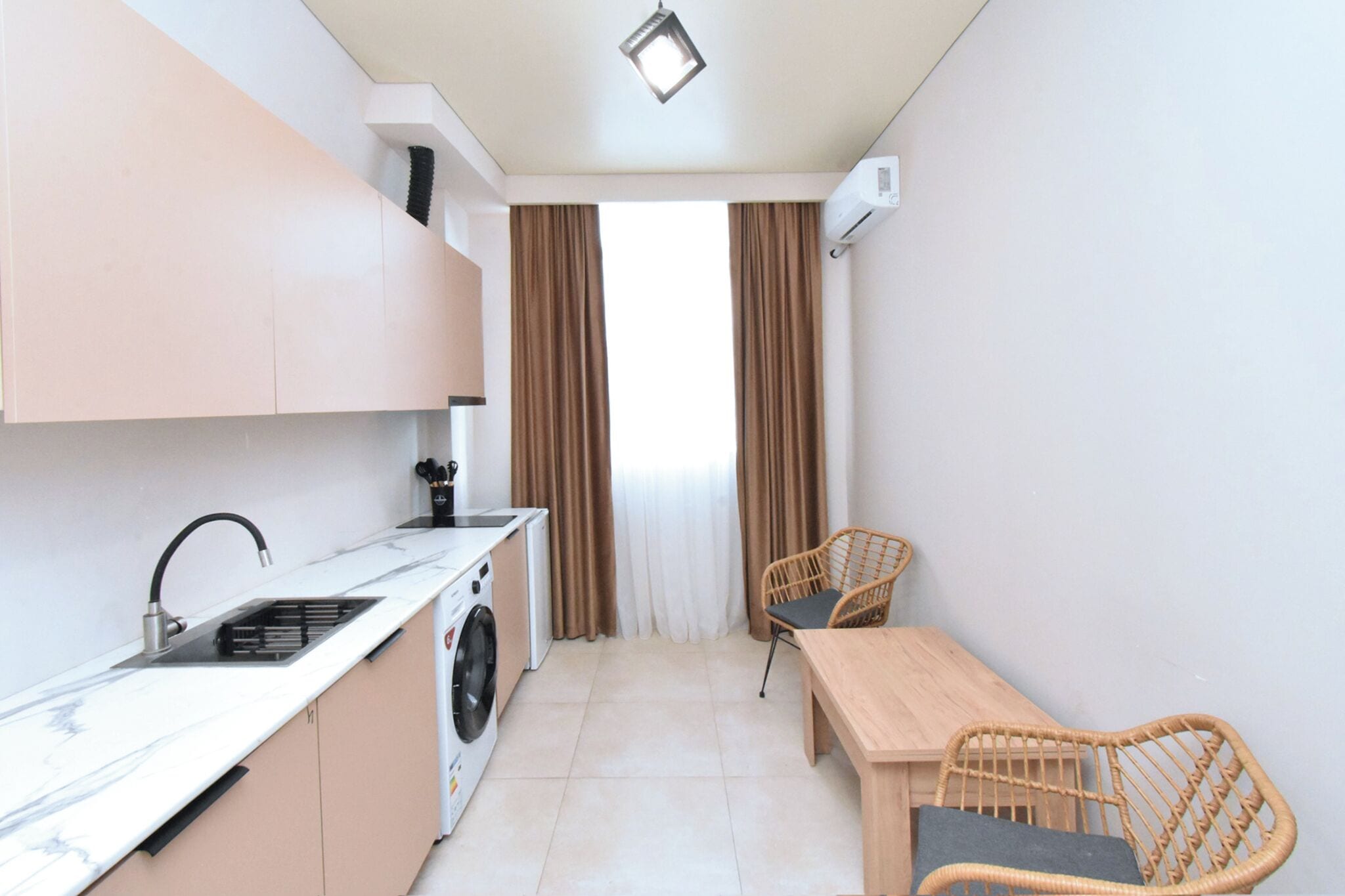 Una cocina compacta con pileta, estufa y lavadora, un comedor con mesa y sillas, y una ventana con cortinas.