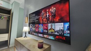 Un televisor de pared muestra varios carteles de películas, incluyendo 'Lucifer' y 'Gotham', en una habitación con un mueble de madera y una lámpara.