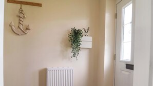 Un pasillo con un radiador blanco, un adorno de ancla colgante, una planta en maceta sobre un estante blanco y una puerta con ventanal.