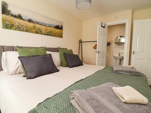 Una cama bien hecha con almohadas, una manta verde y toallas dobladas. En la pared hay un cuadro de un campo, y se ve un baño a través de una puerta abierta.