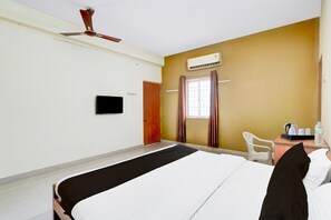 Quarto de hotel com cama, escrivaninha com cadeira, televisão, ar condicionado e janela com cortinas.