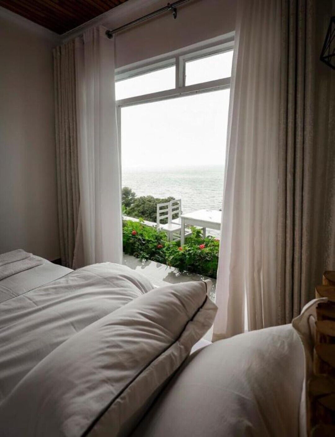 A bedroom with a bed, white curtains, and a view of the sea through a window.