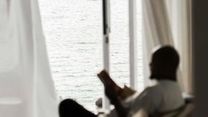 A room with a large window, a person sitting on a chair, and a view of the sea.
