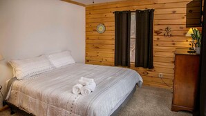 Ein Schlafzimmer mit einer Holzwand, einem Bett mit gemusterten Textilien, einer Uhr und einem Fenster mit Vorhängen.