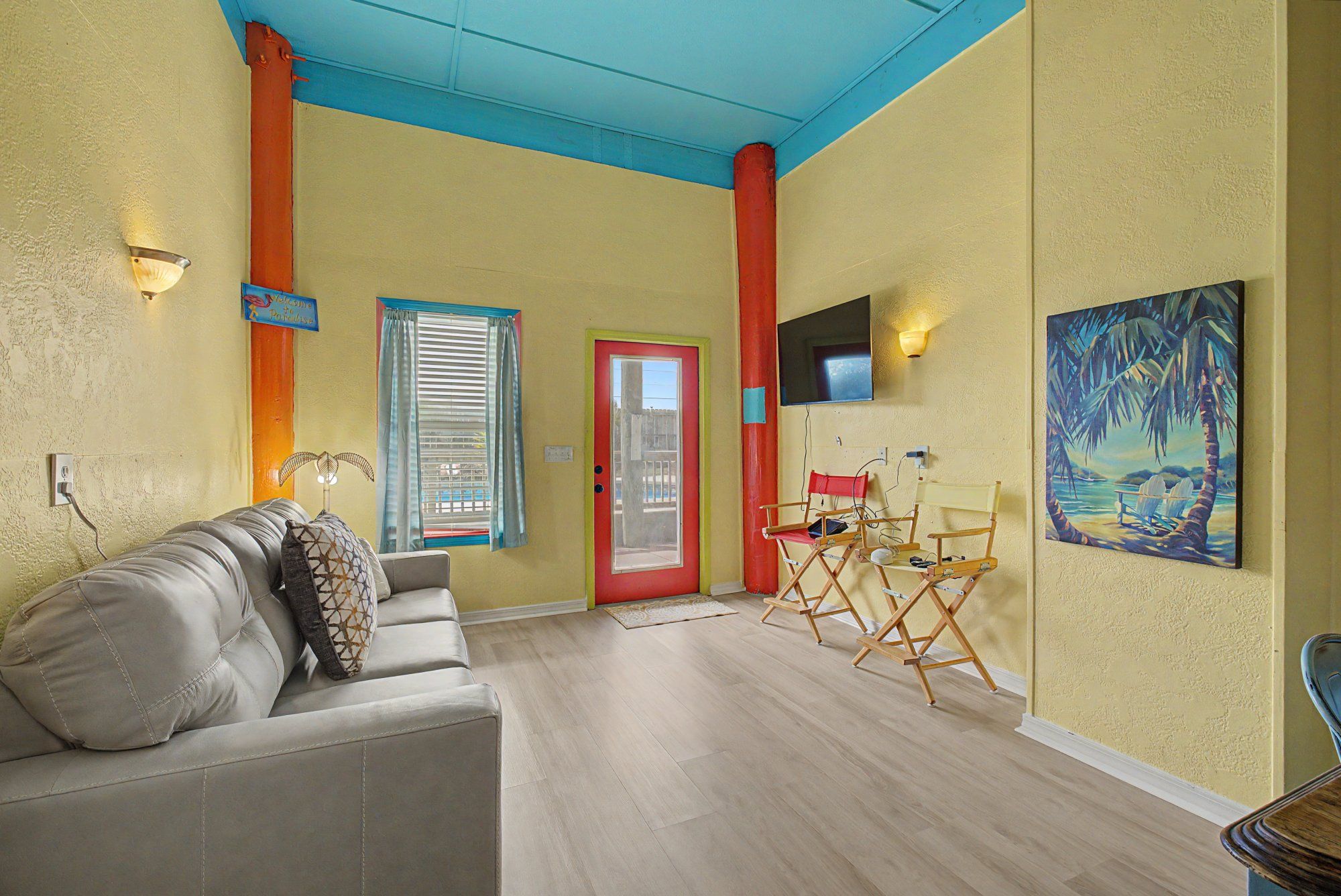 Una sala de estar con un sofá, una puerta roja, una ventana con cortinas, un cuadro y un televisor en la pared.