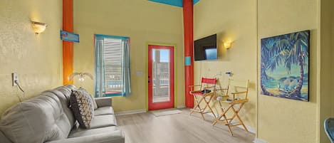 Una sala de estar con un sofá, una puerta roja, una ventana con cortinas, un cuadro y un televisor en la pared.
