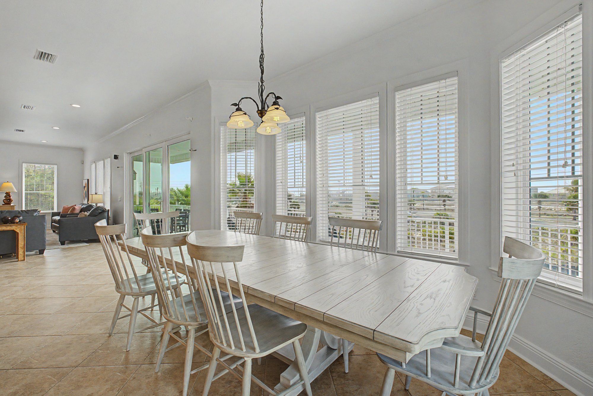 Un vaste espace dédié à la salle à manger, avec une longue table en bois, des chaises blanches et de grandes fenêtres équipées de stores.
