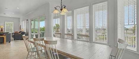 Un vaste espace dédié à la salle à manger, avec une longue table en bois, des chaises blanches et de grandes fenêtres équipées de stores.