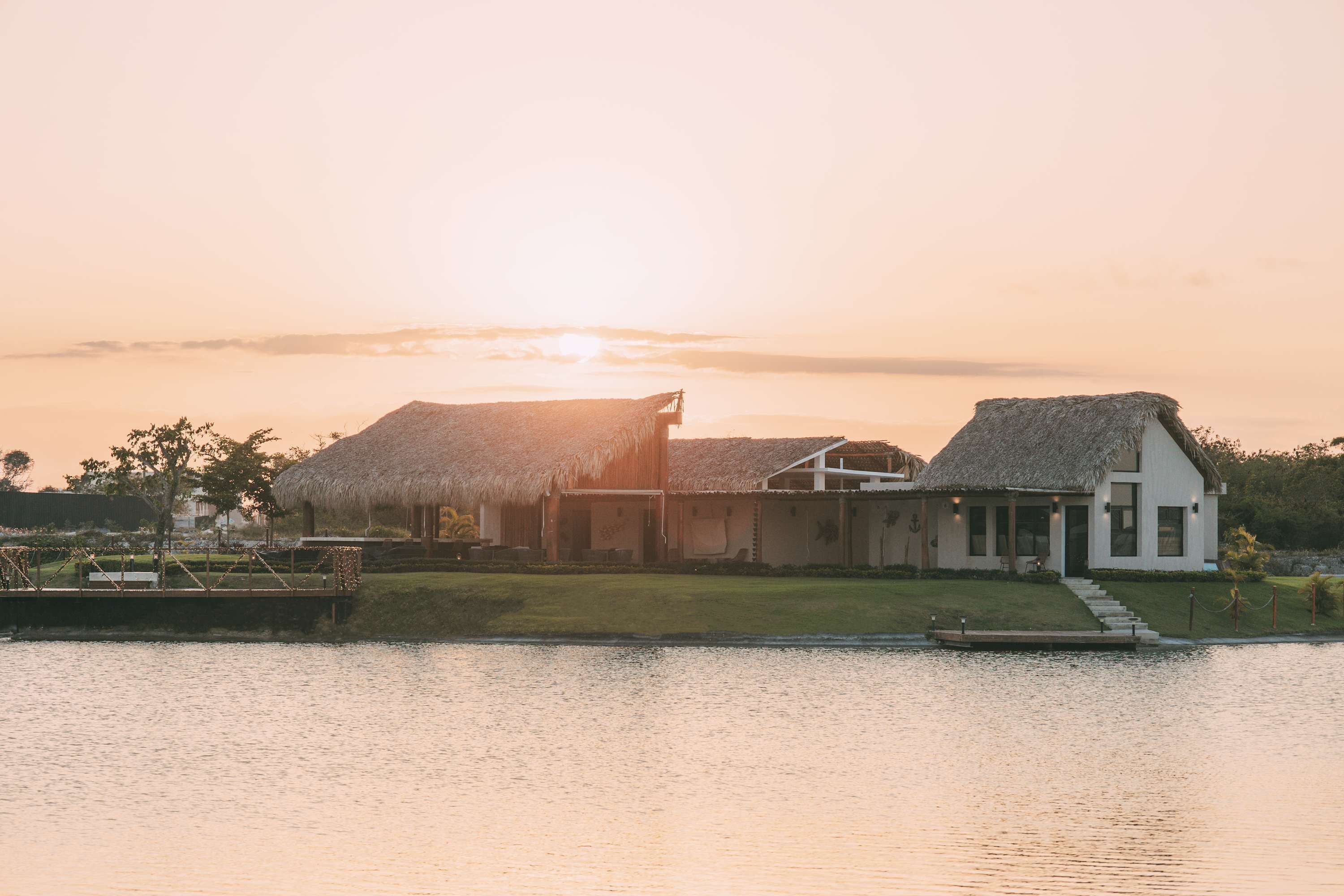 A house with a thatched roof and a modern extension by a lake at sunset.