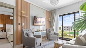 A modern living room with a TV, two armchairs, a sofa, and a large window with a view of buildings. - NOK Modern 2BR With Rooftop Jacuzzi By Vistacana (Punta Cana)
