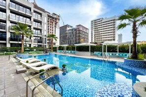 Ein Swimmingpool mit Liegestühlen, einer überdachten Sitzgruppe und modernen Gebäuden im Hintergrund.