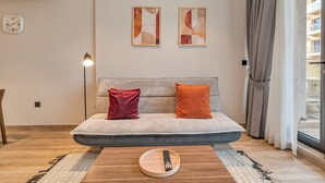 Uma sala de estar moderna com sofá, controle remoto em um suporte, arte abstrata na parede e uma janela com cortinas.