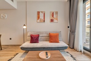 A modern living room with a sofa, a remote control on a tray, abstract art on the wall, and a window with curtains. - Free Housekeeping Visits During Your Stay - Stayshort - Sophisticated 1 Bedroom Apartment in JVC That Sleeps 4 (Dubai)