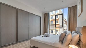Quarto de hotel com uma cama grande, um guarda-roupa, uma janela com vista para prédios e um abajur.