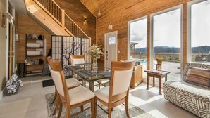 A spacious living area with a wooden staircase, a dining table with chairs, a sofa, and large windows offering a view of the outdoors. - Poolside ADU w Sauna Free Tix Tiki Bar Retreat (Candler)