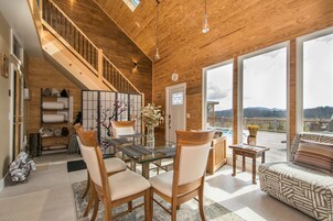 Uma sala de estar ampla com escada de madeira, mesa de jantar com cadeiras, sofá e grandes janelas com vista para o exterior.