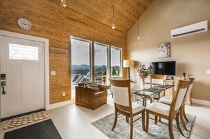 Uma sala de estar moderna com teto de madeira, área de jantar com mesas de vidro e uma grande janela com vista para as montanhas.