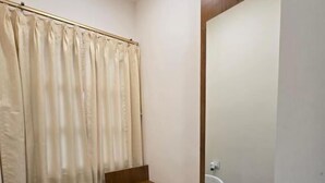 A small bathroom with a wooden vanity, a white chair, a mirror, and a beige curtain. - Vaikuntha Stays Hotel (Indore)