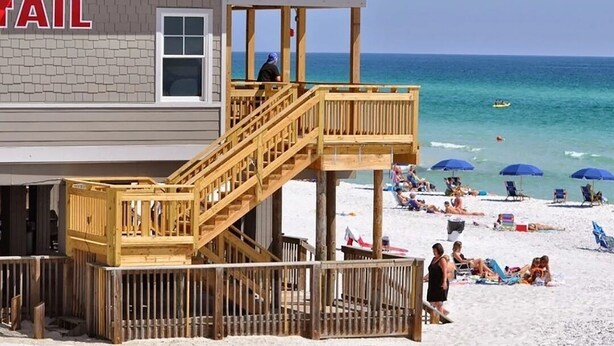 A beachside building with a wooden deck, a lifeguard tower, and a beach with umbrellas and people. - Your Front-Row Seat to Paradise, Pool, Balconies – Sleeps 14 (Miramar Beach)