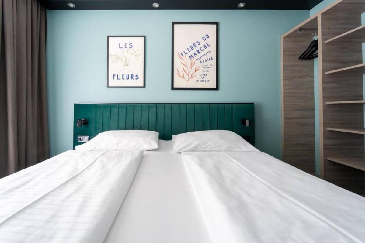 Een tweepersoonsbed met witte lakens, een blauwgroen hoofdbord en twee ingelijste kunstwerken aan de muur.