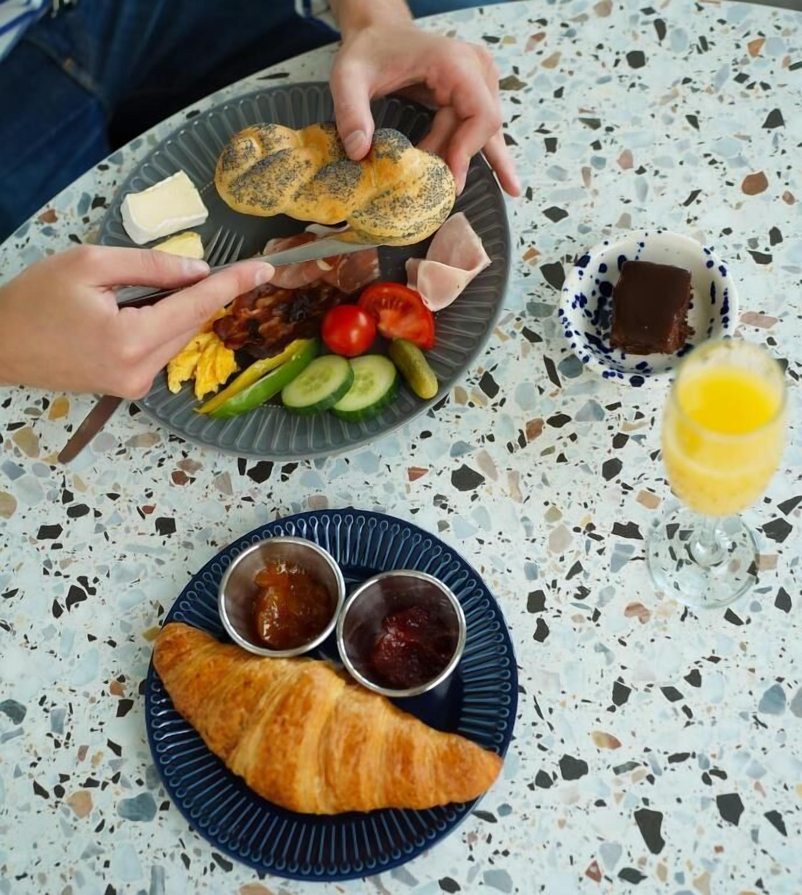 A table with a plate of food including bread, eggs, tomatoes, cucumbers, and a croissant, along with condiments and a glass of juice.