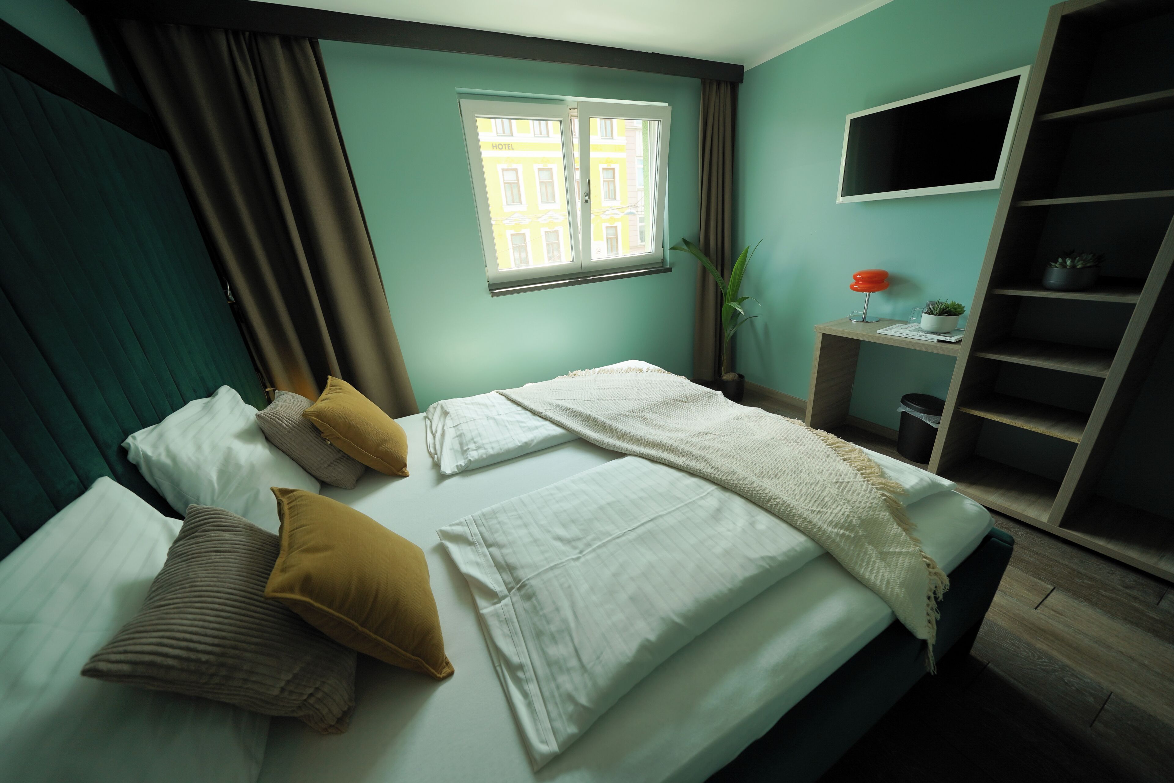 Habitación de hotel con cama, ventana con cortinas, una planta, escritorio con lámpara y estantería.