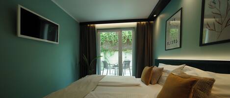 A hotel room with a bed, a TV, a balcony with chairs, and a view of greenery.