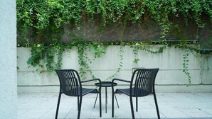 Eine Terrasse mit zwei schwarzen Metallstühlen und einem kleinen runden Tisch, umgeben von einer Betonmauer, die mit Grünspan überwuchert ist.