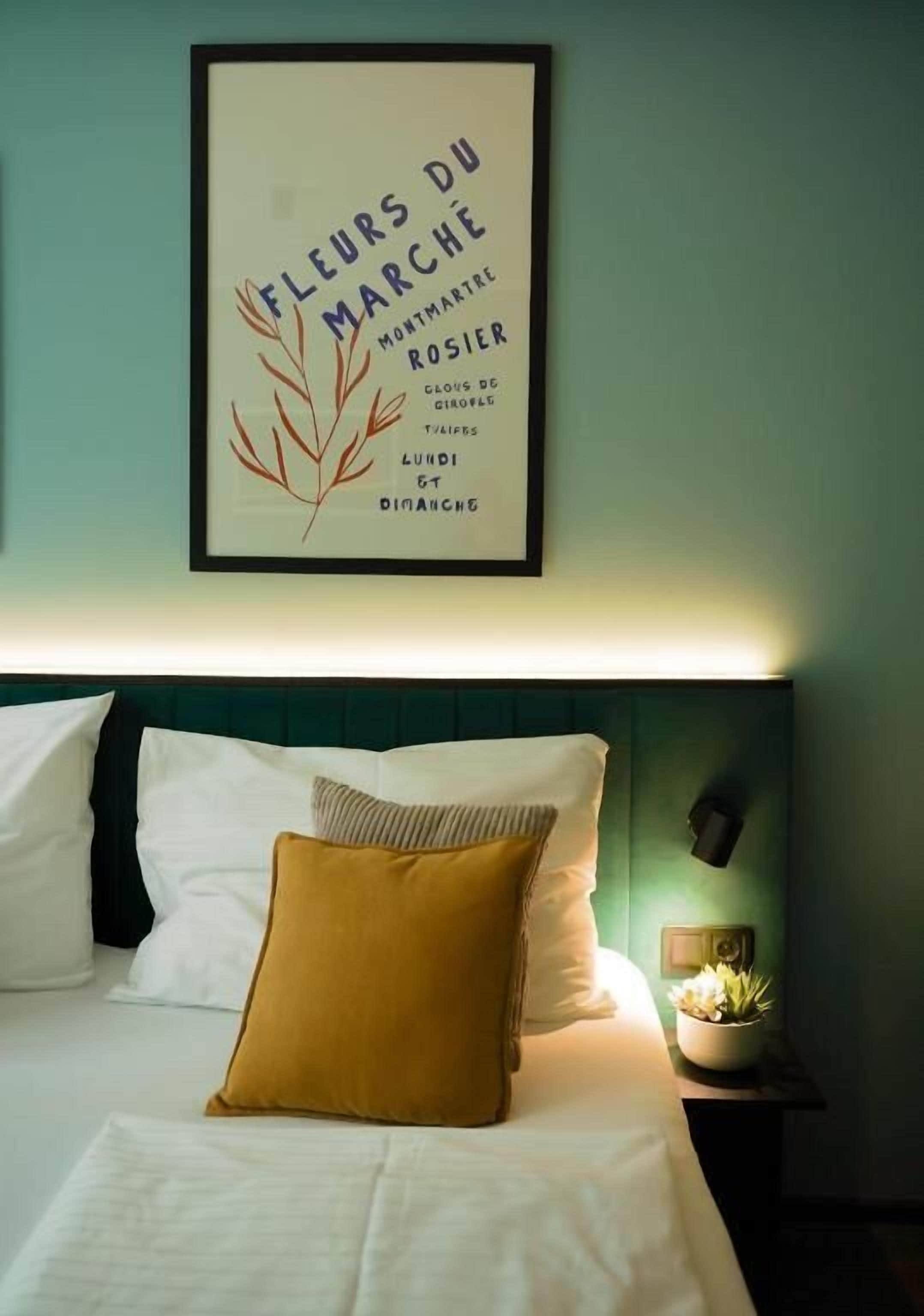 Un letto rifatto con cuscini bianchi e color senape, un quadro appeso al muro e una piccola pianta sul comodino.