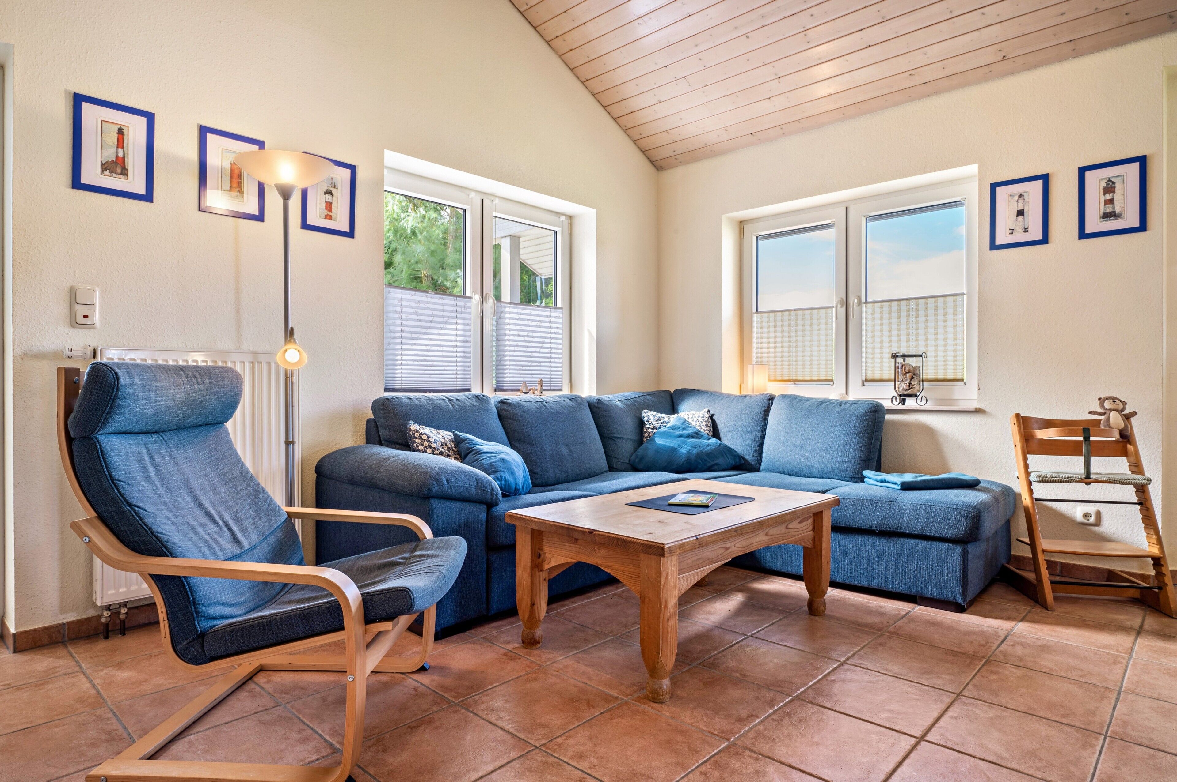 Una sala de estar con un sofá azul, una mesa de centro de madera y un sillón mecedor. Hay dos ventanas con persianas, y una lámpara en el suelo.