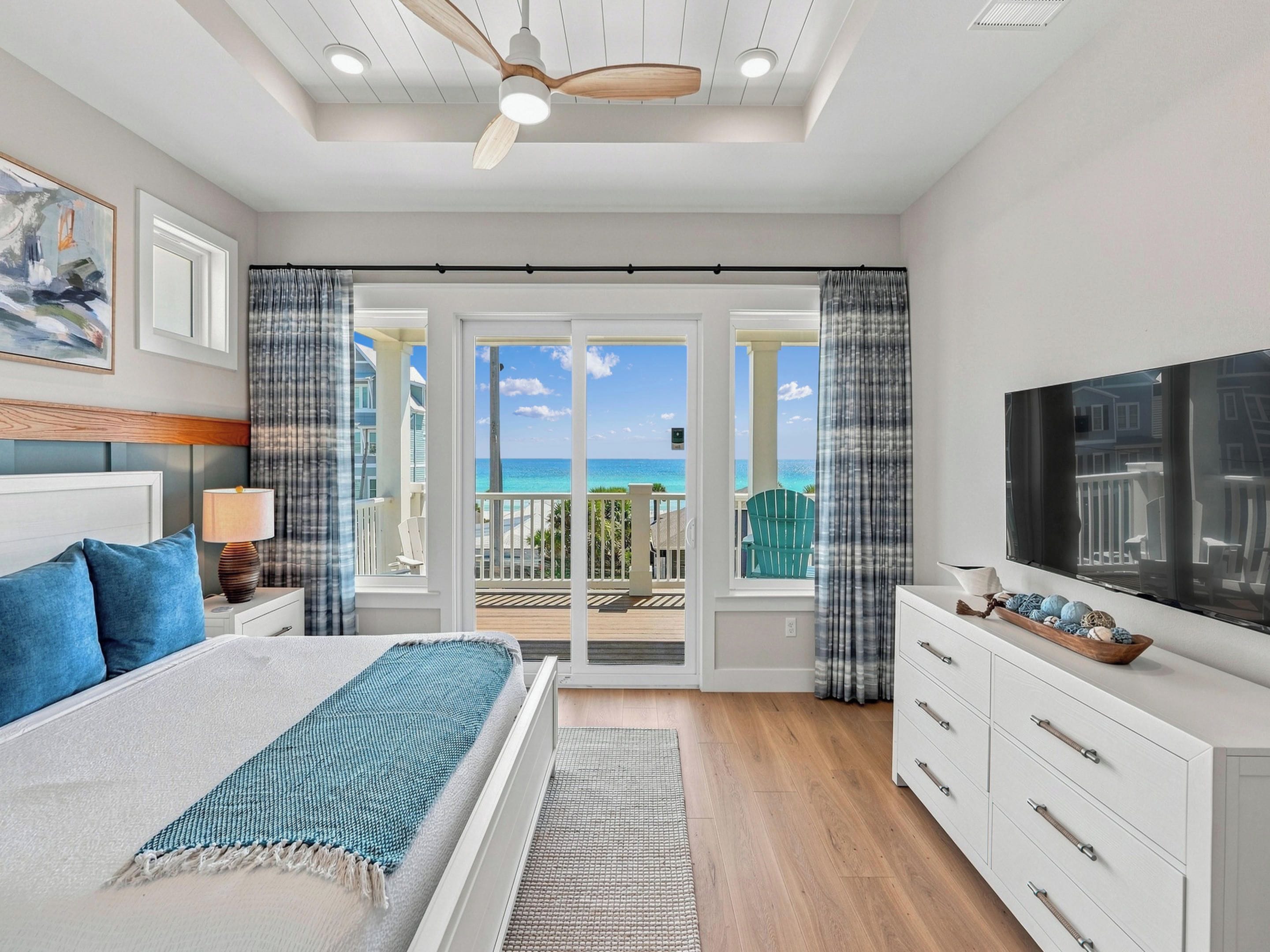 A bedroom with a bed, a TV, a dresser, and a balcony with a view of the ocean.