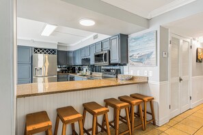 A kitchen with a central island, bar stools, and a view of the adjacent dining area. - Summit Penthouse 3BR 3BA, Great Location! (Panama City Beach)