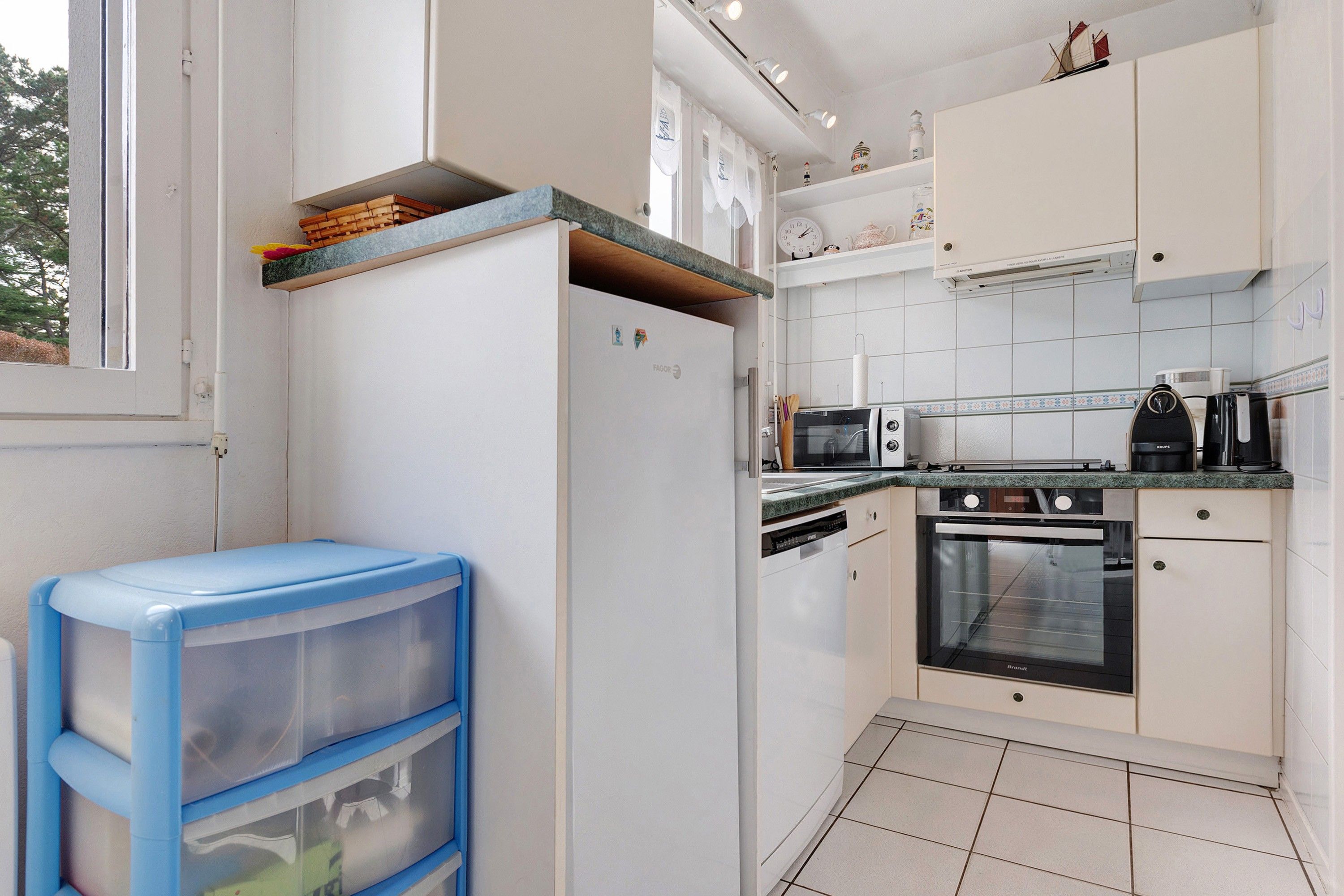 A compact kitchen with white cabinets, a built-in oven, microwave, and a dishwasher. There is a small shelf with decorative items and a window with a view of trees.
