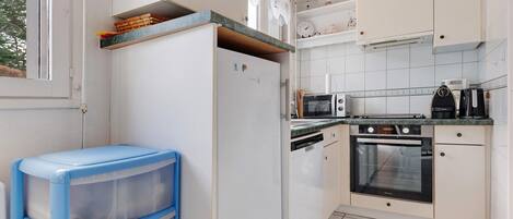 A compact kitchen with white cabinets, a built-in oven, microwave, and a dishwasher. There is a small shelf with decorative items and a window with a view of trees.