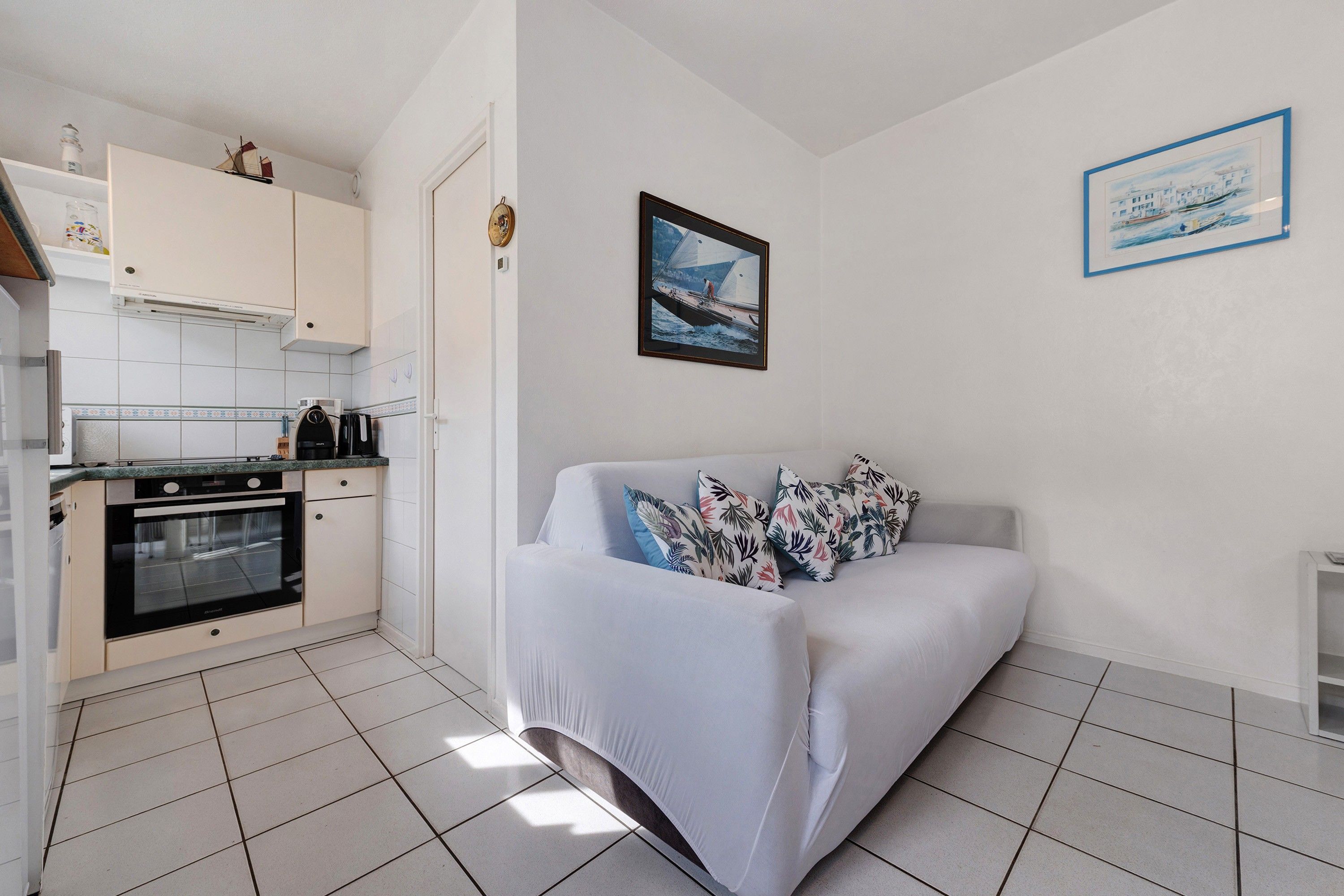 A compact living space with a kitchenette, a sofa with patterned cushions, and two framed artworks on the walls.
