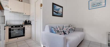 A compact living space with a kitchenette, a sofa with patterned cushions, and two framed artworks on the walls.