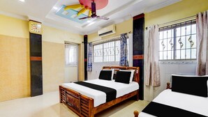 Quarto de hotel com duas camas, um ventilador de teto, aparelho de ar condicionado, janela com grades de metal e um relógio de parede.