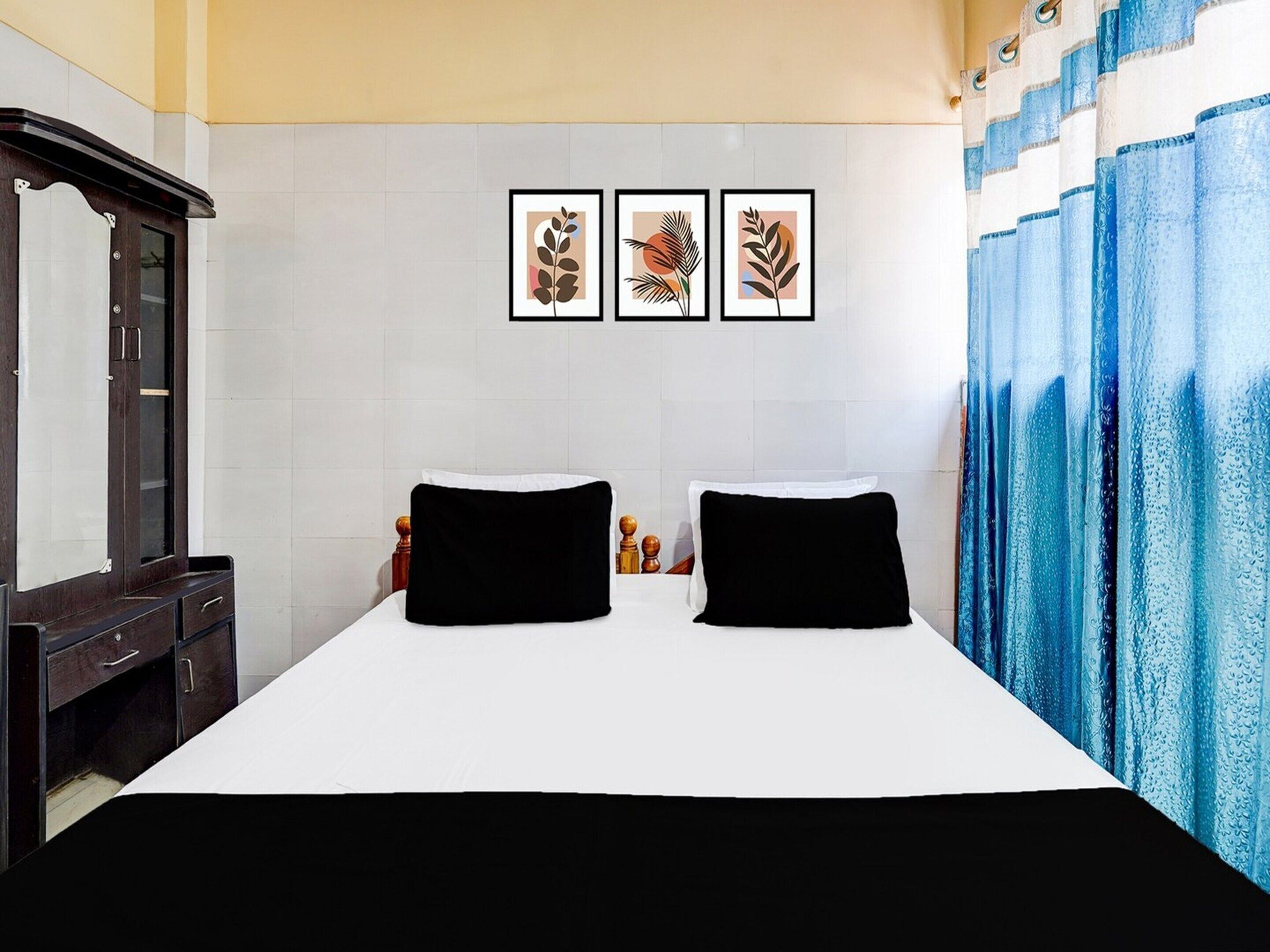 Ein Hotelzimmer mit einem Bett, zwei schwarzen Kissen, einer Holzkommode, einem Spiegel und drei gerahmten Pflanzenbildern an der Wand.