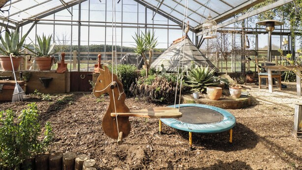 Infern de vidre amb un cavall de fusta com a cadira, un trampolí i diverses plantes en testos.