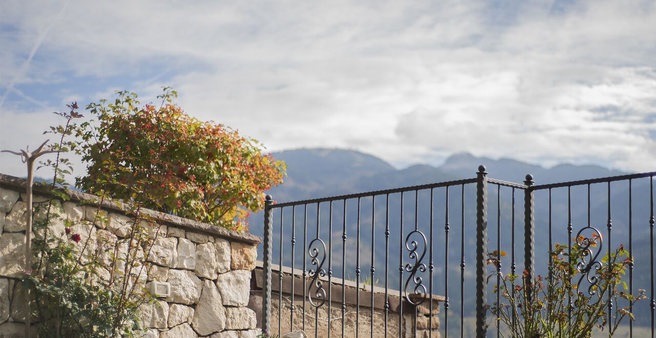 Un muro de piedra con una reja de metal, una planta floreciente y montañas al fondo.