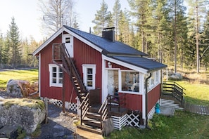 A red wooden cabin with a staircase, a porch, and a view of a forest.